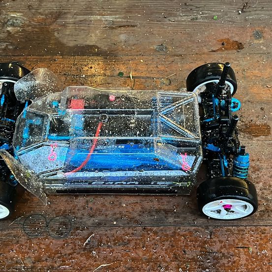 Ein blaues und schwarzes ferngesteuertes Auto mit durchsichtigem Gehäuse liegt auf einer Holzoberfläche und zeigt Abnutzungserscheinungen.
