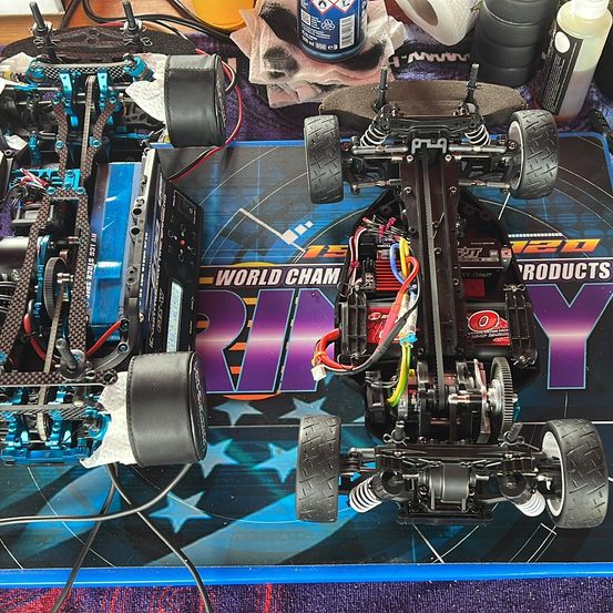 Ein RC-Auto liegt auf einem blauen Teppich mit den Worten 'World Cham RIP Y'. Drähte und andere Komponenten sind sichtbar. Flaschen und Werkzeuge sind herumgestreut.