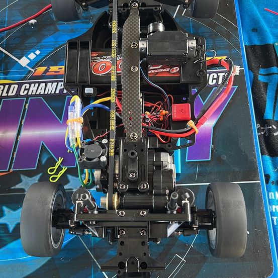 Eine Nahaufnahme eines RC-Autos auf einem blau-lila Hintergrund mit der Aufschrift 'World Champion'. Das Auto hat ein schwarzes Chassis, sichtbare Kabel und mechanische Komponenten.
