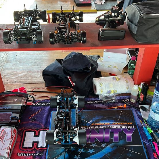 Ein Arbeitstisch zeigt mehrere ferngesteuerte Fahrzeuge, darunter eines auf einem Tuch mit dem Wort 'H'. In der Nähe befinden sich ein Werkzeugkasten, Werkzeuge und verschiedene Flaschen. Auf einem Regal stehen weitere Fahrzeuge, eine Kamera und Taschen.