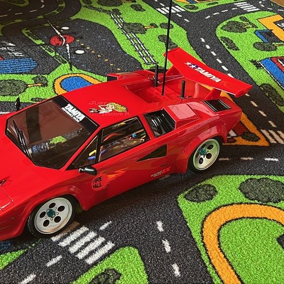 Ein rotes Lamborghini Modellauto steht auf einem grünen und schwarzen Spielteppich mit Straßenmarkierungen. Das Auto hat einen großen Flügel hinten und die Aufschrift 'FIRE BALL' an der Seite.