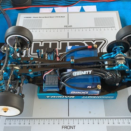 Ein blaues Tamiya-Modellauto liegt auf einem blauen Tuch mit einem Lineal, das seine Länge misst. Es hat ein schwarzes und blaues Chassis, mit verschiedenen Komponenten wie Batterie, Motor und Zahnrädern sichtbar.