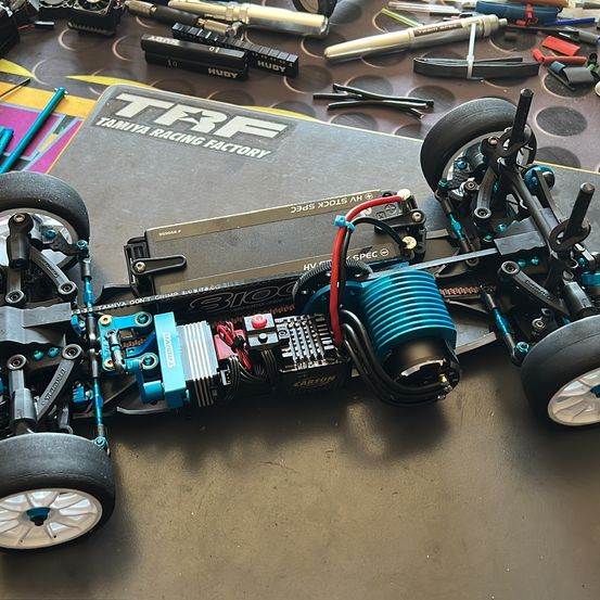 Ein Tamiya-Rennwagen-Chassis mit freiliegenden Elektronikbauteilen steht auf einer Arbeitsfläche. Das Chassis hat vier Räder, einen Akku und verschiedene Kabel und Komponenten.