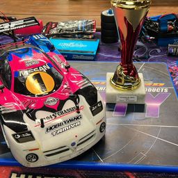 Ein rosa und weißer Rennwagen mit Hobbywing Tamiya-Marke steht neben einem rot-goldenen Pokal auf einer Streckenlayoutmatte.
