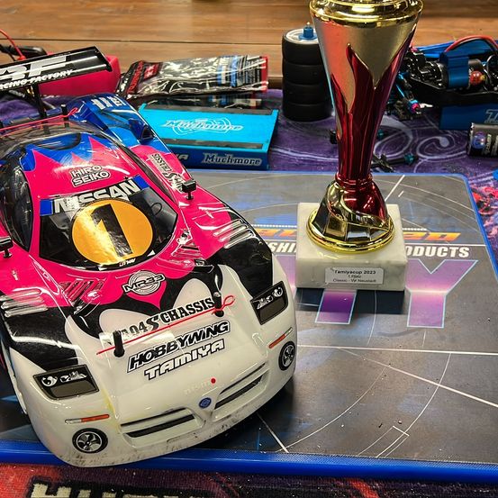 Ein rosa und weißer Rennwagen mit Hobbywing Tamiya-Marke steht neben einem rot-goldenen Pokal auf einer Streckenlayoutmatte.