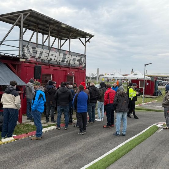 Eine Gruppe von Menschen in Winterkleidung steht vor einem roten Gebäude mit der Aufschrift Team RCC. Einige tragen Mützen und Sneakers, andere Handschuhe. Sie stehen auf einer Straße in der Nähe weißer Linien und eines Grasbereichs.
