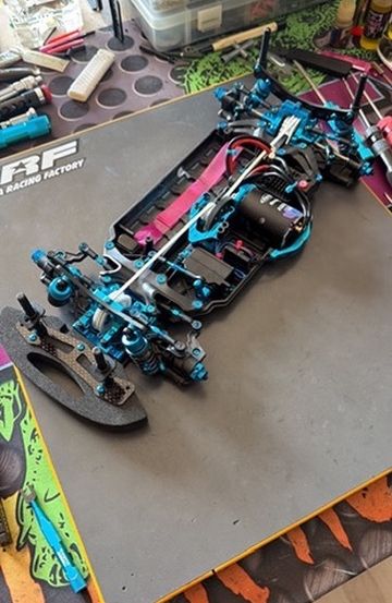 Ein blaues und schwarzes RC-Auto-Chassis liegt auf einem grauen Teppich mit einem Racing-Factory-Logo. Verschiedene Werkzeuge und Teile sind darauf verstreut auf einem bunten Tischtuch.