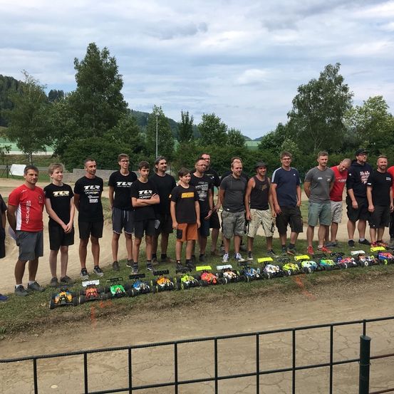 Eine Gruppe von Menschen steht neben einer Rennstrecke mit Spielzeugautos, in Freizeitkleidung, mit Bäumen und Bergen im Hintergrund.