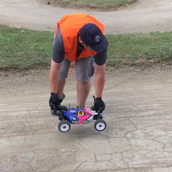 Ein Mann in einer orangefarbenen Weste und einem Hut beugt sich herunter, um ein kleines ferngesteuertes Auto auf einer Erdstrecke zu untersuchen.