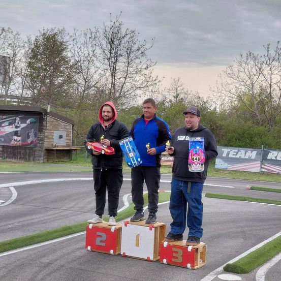 Drei Männer stehen auf Podiumsstufen und halten Trophäen und Modellautos. Der Ort ist eine Rennstrecke mit einem Grasbereich und Bäumen.