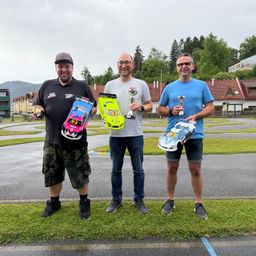 Drei Männer posieren mit Trophäen und Spielzeugautos auf einer Rennstrecke. Hinter ihnen ist ein Gebäude mit rotem Dach zu sehen, und Bäume säumen den Hintergrund.