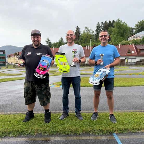 Drei Männer posieren mit Trophäen und Spielzeugautos auf einer Rennstrecke. Hinter ihnen ist ein Gebäude mit rotem Dach zu sehen, und Bäume säumen den Hintergrund.