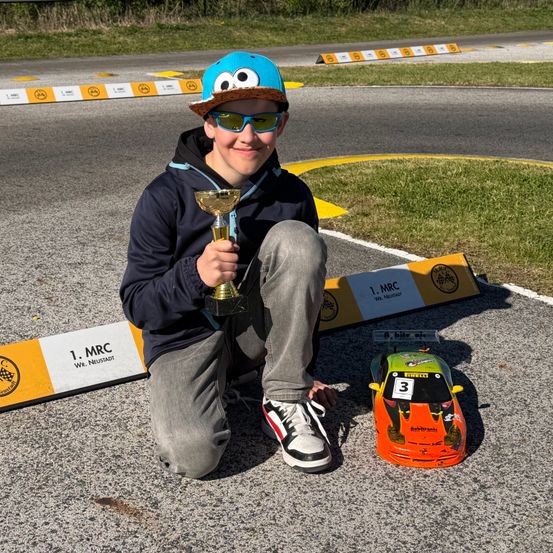 Ein Junge mit blauer Mütze und Sonnenbrille hält einen Pokal, während er auf der Rennstrecke kniet. Neben ihm steht ein Spielzeugauto mit der Nummer 3.