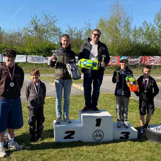 Sechs junge Rennfahrer stehen auf einem Podium mit drei Stufen, halten Trophäen und Autos. Sie tragen Medaillen um den Hals. Das Podium ist mit 1, 2 und 3 nummeriert.