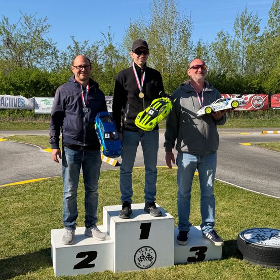 Drei Männer stehen auf einem Podium auf einer Rennstrecke, jeder hält ein ferngesteuertes Auto. Der Mann auf der linken Seite hat eine Silbermedaille, die Mitte eine Goldmedaille und der rechte eine Silbermedaille. Die Podeste sind mit 1, 2 und 3 nummeriert.
