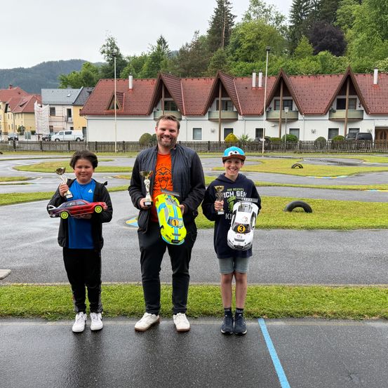 Drei Menschen stehen auf einer nassen Straße und halten Trophäen. Sie stehen vor einer Reihe von Häusern mit roten Dächern. Die beiden Jungen tragen Turnschuhe und halten Spielzeugautos. Der Mann trägt eine Jacke und einen Hut.