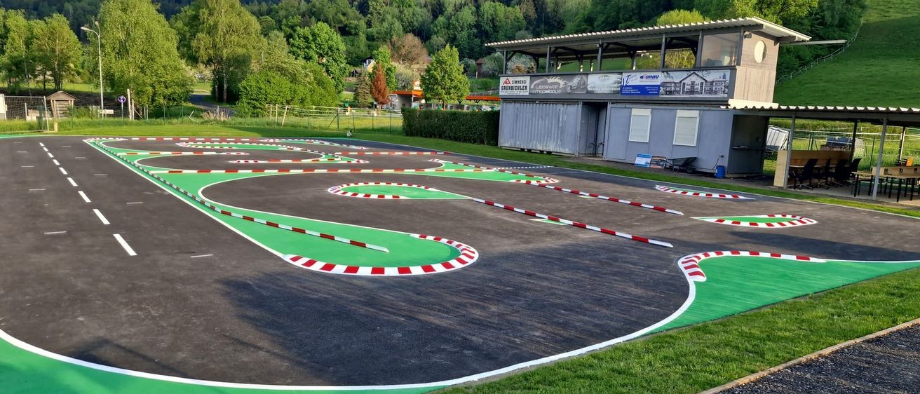 Eine Rennstrecke mit grünen und roten Markierungen, umgeben von einem Grasbereich und einem Gebäude mit einem Schild oben. Die Rennstrecke ist leer.