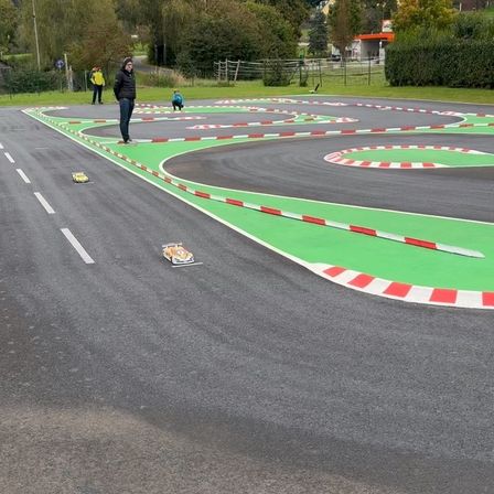Eine Gruppe von Menschen ist auf einer Rennstrecke, wobei eine Person steht und zusieht. Autos fahren auf der Strecke, und zwei Personen sitzen im Gras.