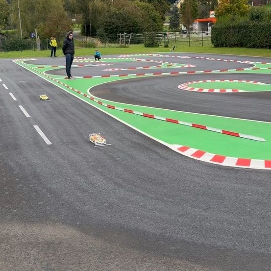 Eine Gruppe von Menschen ist auf einer Rennstrecke, wobei eine Person steht und zusieht. Autos fahren auf der Strecke, und zwei Personen sitzen im Gras.