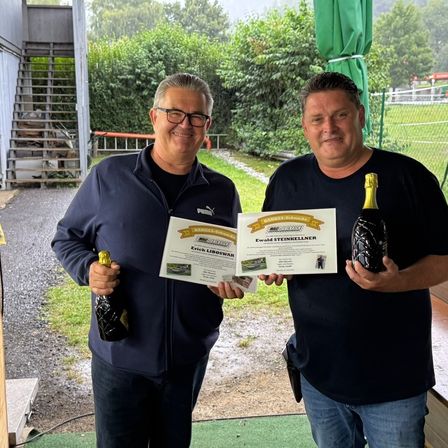 Zwei Männer stehen draußen und halten Champagnerflaschen und Urkunden. Sie lächeln und scheinen zu feiern.