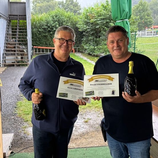 Zwei Männer stehen draußen und halten Champagnerflaschen und Urkunden. Sie lächeln und scheinen zu feiern.