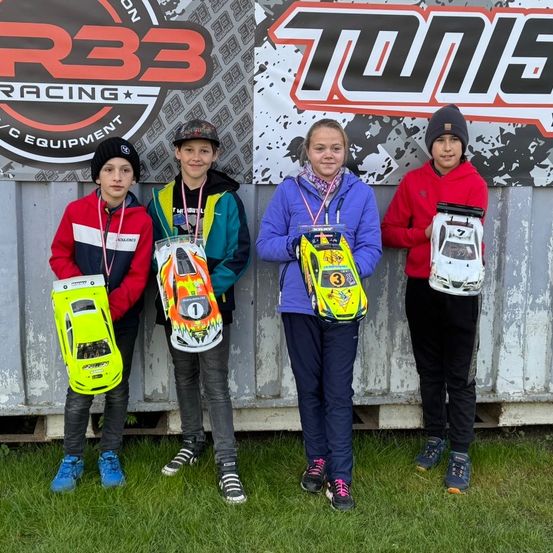 Vier Kinder stehen zusammen und halten ferngesteuerte Autos. Sie lächeln, eines trägt einen Hut. Ein Banner hinter ihnen lautet 'TONE'.