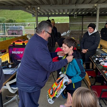 Ein älterer Mann stiftet einem jungen Jungen eine Medaille unter einem Pavillon. Ein Spielzeugauto und ein Banner sind auf dem Tisch hinter ihnen. Menschen stehen und sitzen herum.
