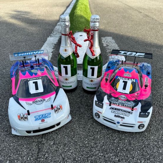 Zwei ferngesteuerte Rennwagen sind mit zwei Champagnerflaschen, die mit '1' beschriftet sind, auf einer Rennstrecke aufgereiht. Ein Auto hat ein pinkes und blaues Design, während das andere weiß ist.