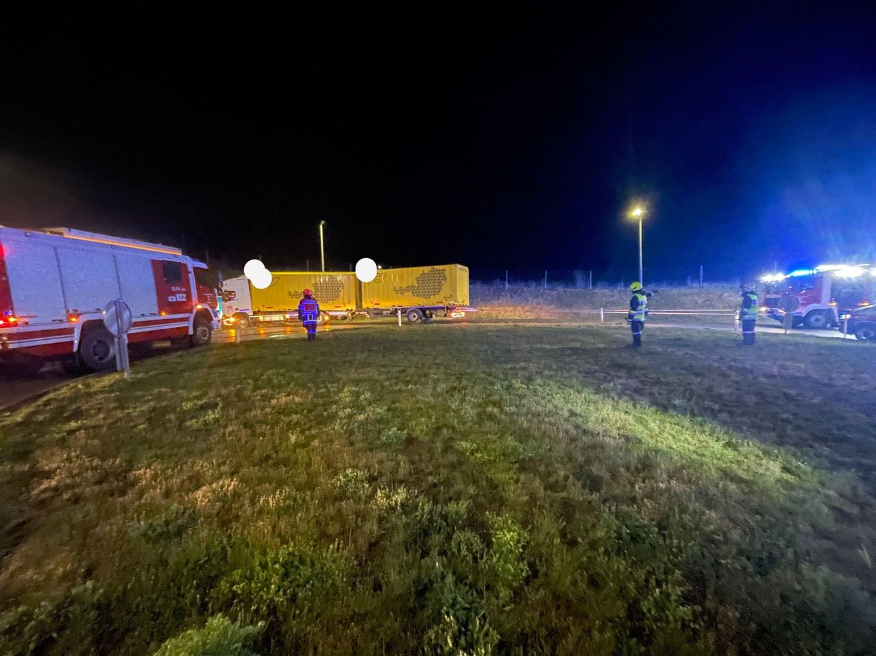 Zwei Notfallhelfer mit gelben Westen stehen neben einem Lkw mit gelber Anhänger auf einem Grasfeld bei Nacht.