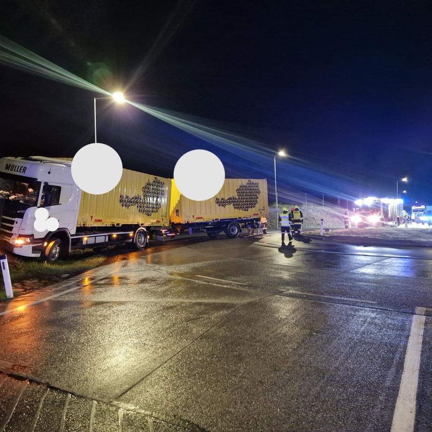 Ein LKW ist nachts auf der nassen Straßenseite geparkt. Eine Person in einer reflektierenden Weste steht in der Nähe.