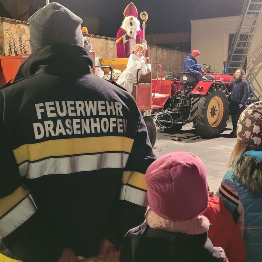 Ein Feuerwehrmann aus Drasenhofen beobachtet eine Parade mit einem Traktor und St. Nikolaus im Hintergrund, während sich Menschen versammeln, um zuzusehen.