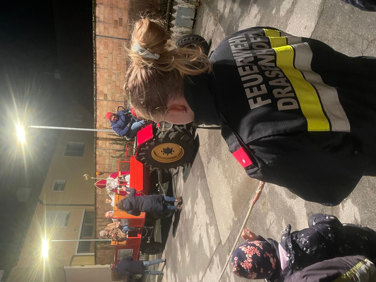 Ein Feuerwehrmann steht hinter einer Menschenmenge, die sich um einen roten Traktor mit einer weißen Figur als Heiliger versammelt. Die Szene spielt sich nachts ab.