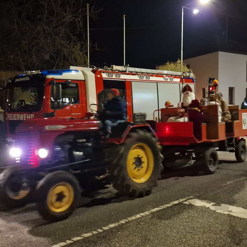 Ein roter Feuerwehrwagen mit einer Weihnachtsmannfigur und Personen auf der Rückseite fährt bei Nacht. Der Fahrer sitzt vorne.