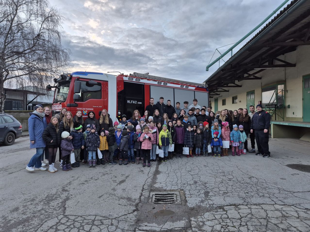 Eine Gruppe von Kindern und Erwachsenen steht zusammen vor einem Feuerwehrauto mit der Aufschrift 'HLFA2 2408'. Einige tragen Mützen und Handschuhe.