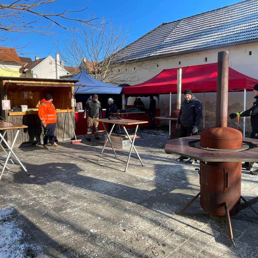 Mehrere Menschen sind an einem sonnigen Tag draußen, mit ein paar Zelten und einem großen roten Zelt. Ein Mann mit einer gelben Jacke steht neben einem Tisch, und ein Mann in einer schwarzen Jacke steht neben einem großen rostigen Ofen.