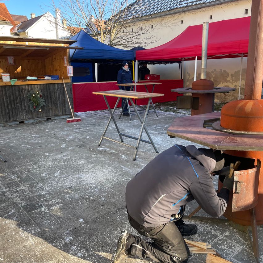 Eine Person kniet neben einem großen Metallofen und fügt Holz hinzu. In der Nähe sind zwei Zelte mit Tischen und einem kleinen Holzständer aufgestellt. Ein Gebäude ist im Hintergrund zu sehen, und der Boden ist verschneit.