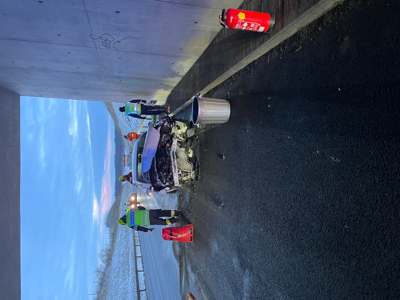 Ein Autounfall auf einer Straße, mehrere Personen in Sicherheitswesten sind anwesend. Ein Feuerlöscher und ein Mülleimer sind in der Nähe.