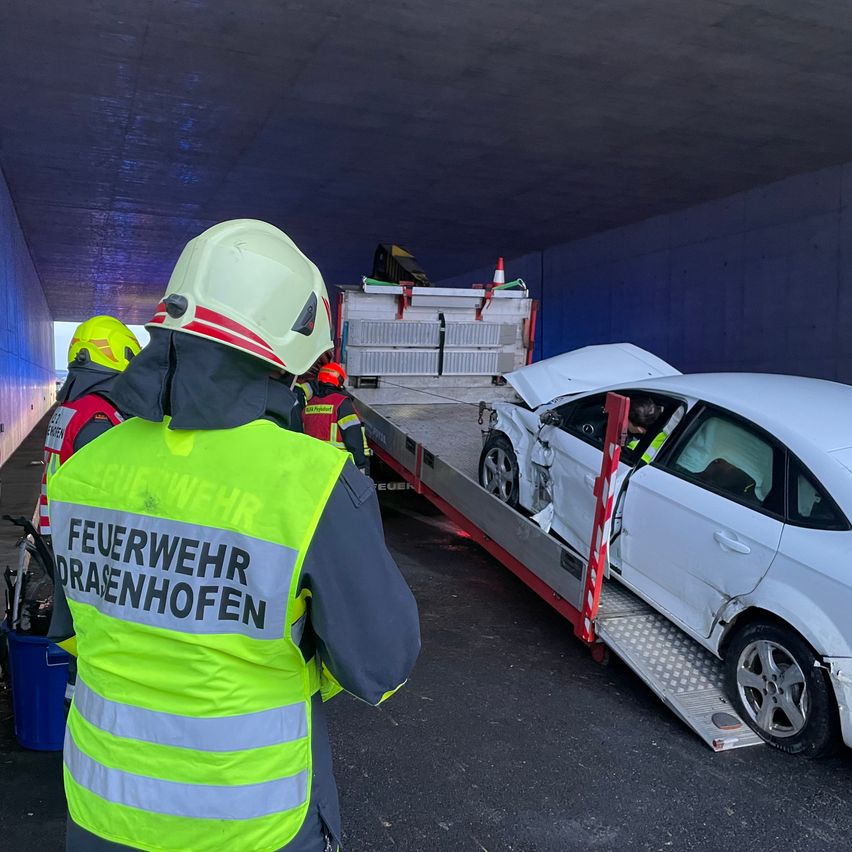 Rettungskräfte entfernen ein beschädigtes weißes Auto aus einem Tunnel. Ein Feuerwehrmann trägt eine reflektierende Weste mit der Aufschrift 'Feuerwehr Drasenhofen.' Das Auto befindet sich auf einem Flachbett-Lkw, dessen Motorhaube geöffnet ist, und in der Nähe steht ein Verkehrskegel.