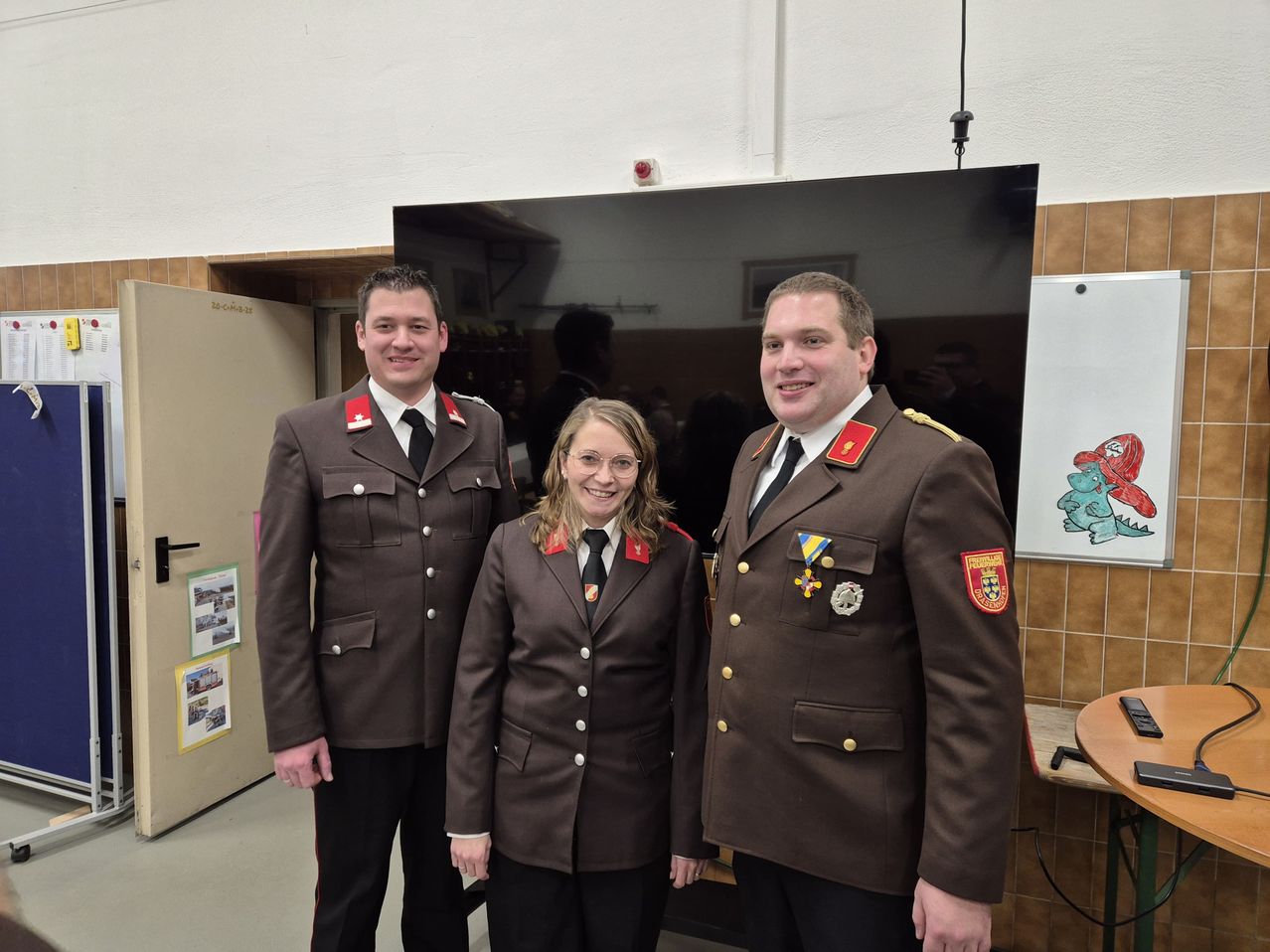 Drei Personen in braunen Uniformen stehen eng nebeneinander vor einem großen Bildschirm. Zwei sind Männer und eine ist eine Frau. Sie alle lächeln für die Kamera.