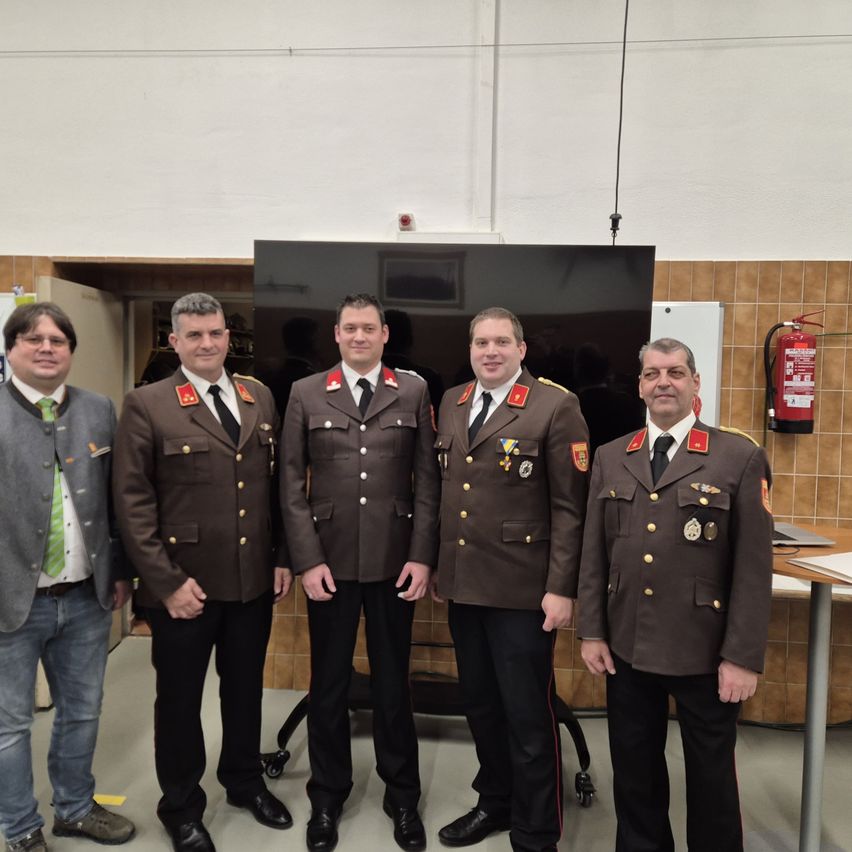 Fünf Männer stehen in einer Reihe in formellen Uniformen; einer trägt eine grüne Krawatte, ein anderer trägt einen grauen Anzug. Sie alle lächeln. Hinter ihnen steht ein Feuerlöscher und ein Monitor.