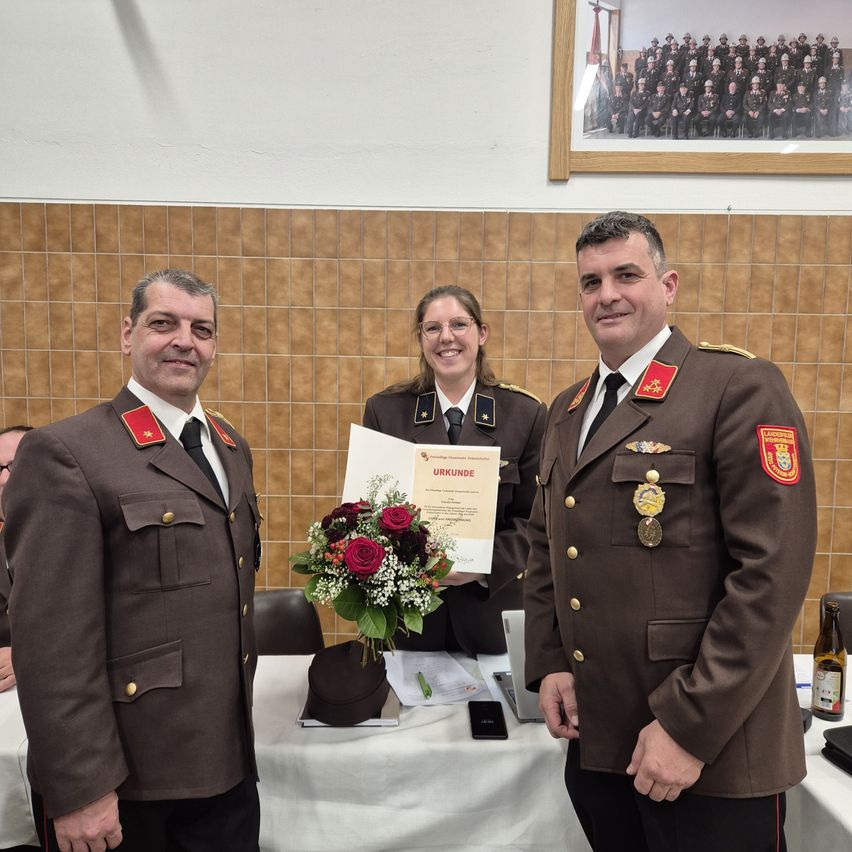 Drei Personen in Militäruniformen stehen lächelnd zusammen. Eine Frau hält ein Zertifikat und einen Blumenstrauß. Hinter ihnen steht ein Tisch mit Laptop, Handy und Flasche. Ein Foto von vielen Leuten in Uniform hängt an der Wand.
