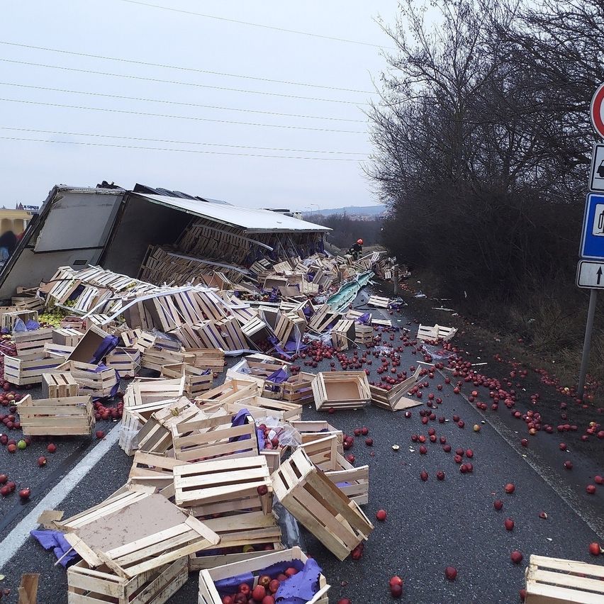 Eine Autobahn ist durch einen umgestürzten LKW blockiert, der seine Ladung roter Äpfel auf die Straße verschüttet. Holzkisten sind verstreut und eine Person mit Helm ist in der Nähe.
