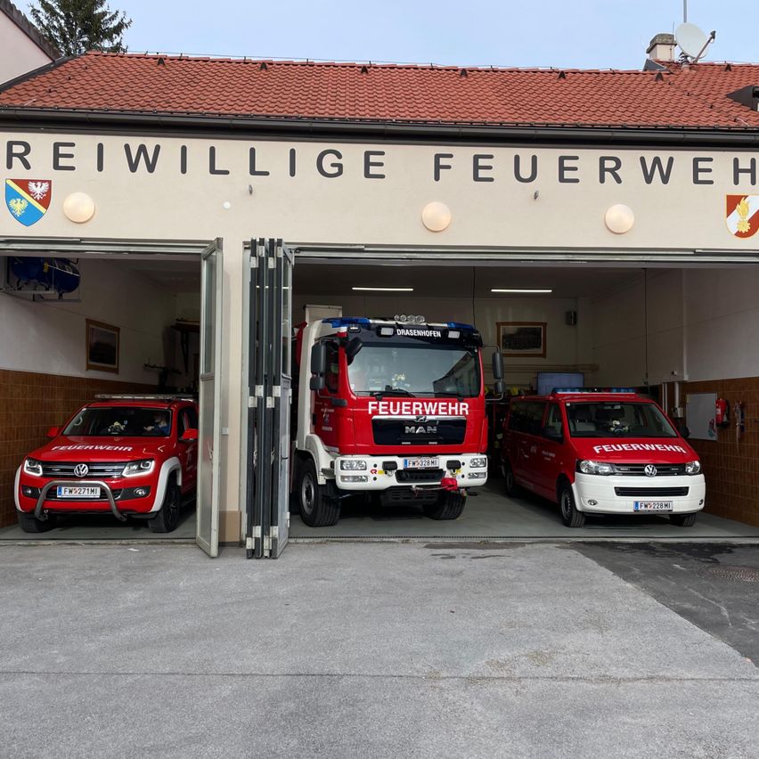Der Eingang einer Feuerwache zeigt drei rote Feuerwehrfahrzeuge und Vans, die im Inneren geparkt sind, mit einem Schild, das 'REIWILLIIGE FEUERWEHR' über dem Eingang steht.