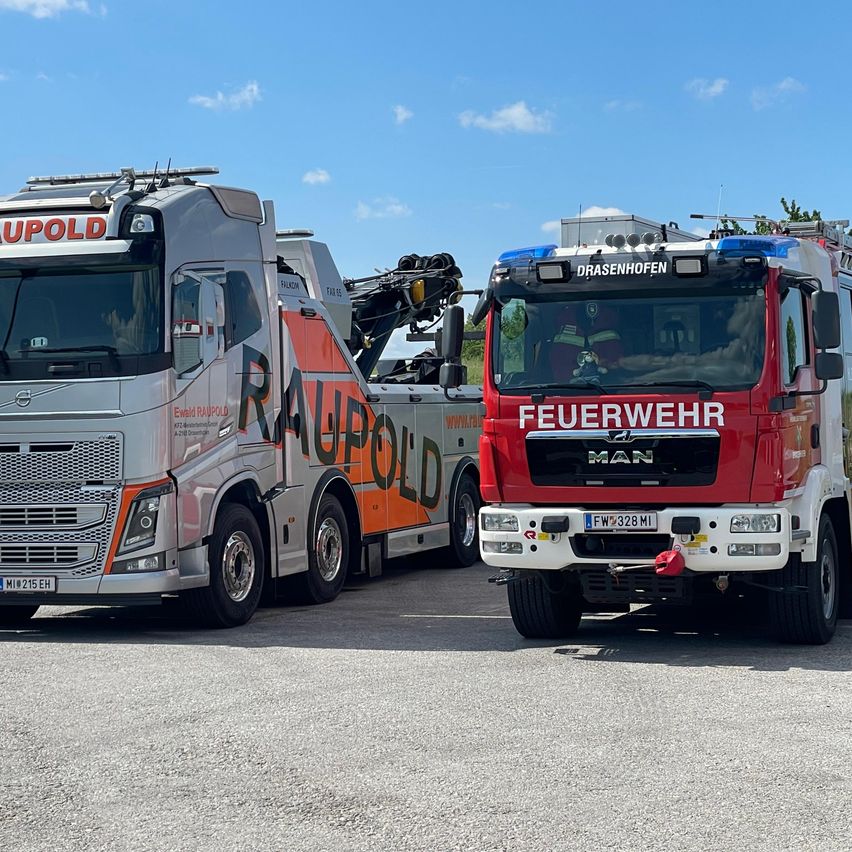 Zwei Lkws sind auf einem Parkplatz geparkt. Der erste Lkw ist weiß und orange, mit der Aufschrift 'Raupold'. Der zweite Lkw ist rot, mit der Aufschrift 'Feuerwehr', und ein Feuerwehrmann sitzt im Inneren.