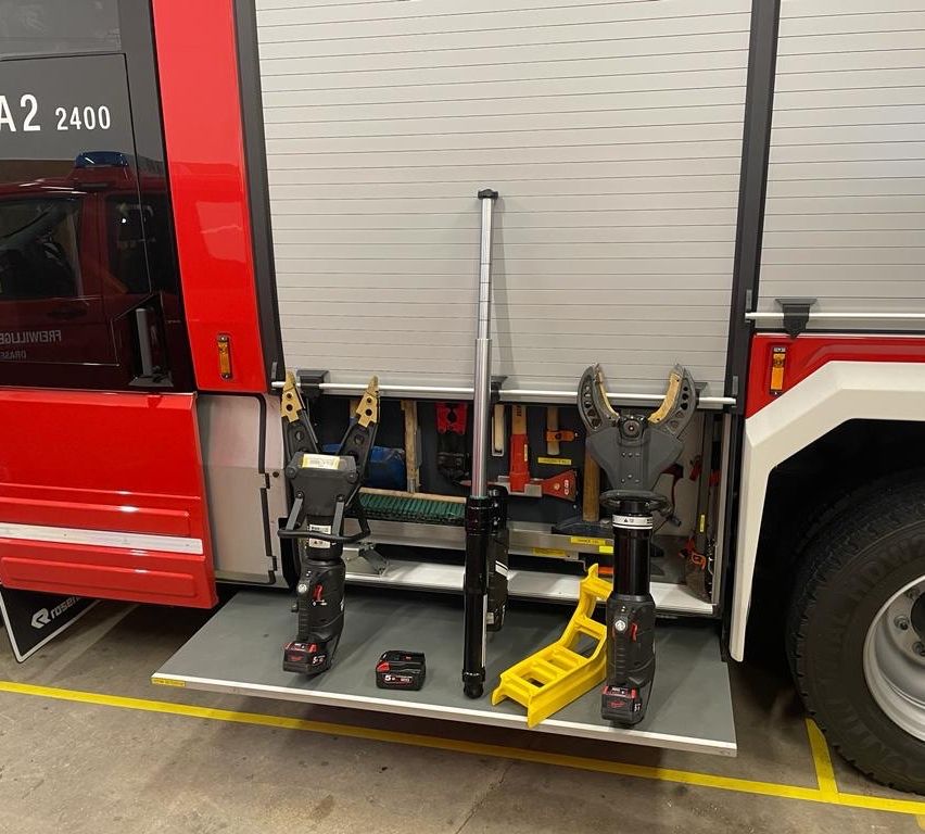 Feuerwehrfahrzeug mit offener Hecktür, zeigt Werkzeuge und Ausrüstung. Eine gelbe Leiter ist auf der Rampe platziert. Das Fahrzeug hat die Nummer 2400 an der Tür.