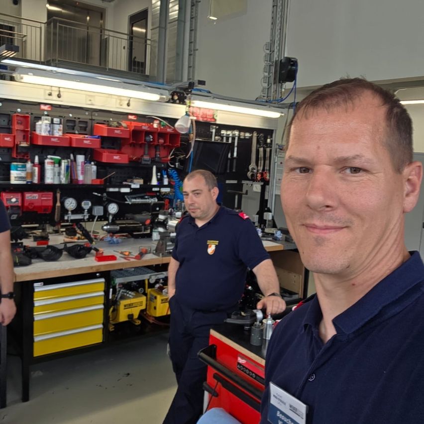 Zwei Männer in blauen Uniformen arbeiten in einer Garage. Hinter ihnen steht ein Arbeitstisch mit verschiedenen Werkzeugen. Der Raum ist mit Deckenleuchten gut beleuchtet.
