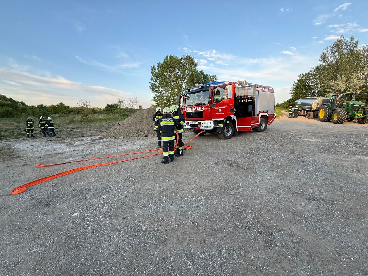 Feuerwehrleute in Uniform verbinden Schläuche mit einem Feuerwehrauto auf einem Kiesweg unter einem teilweise bewölkten Himmel.