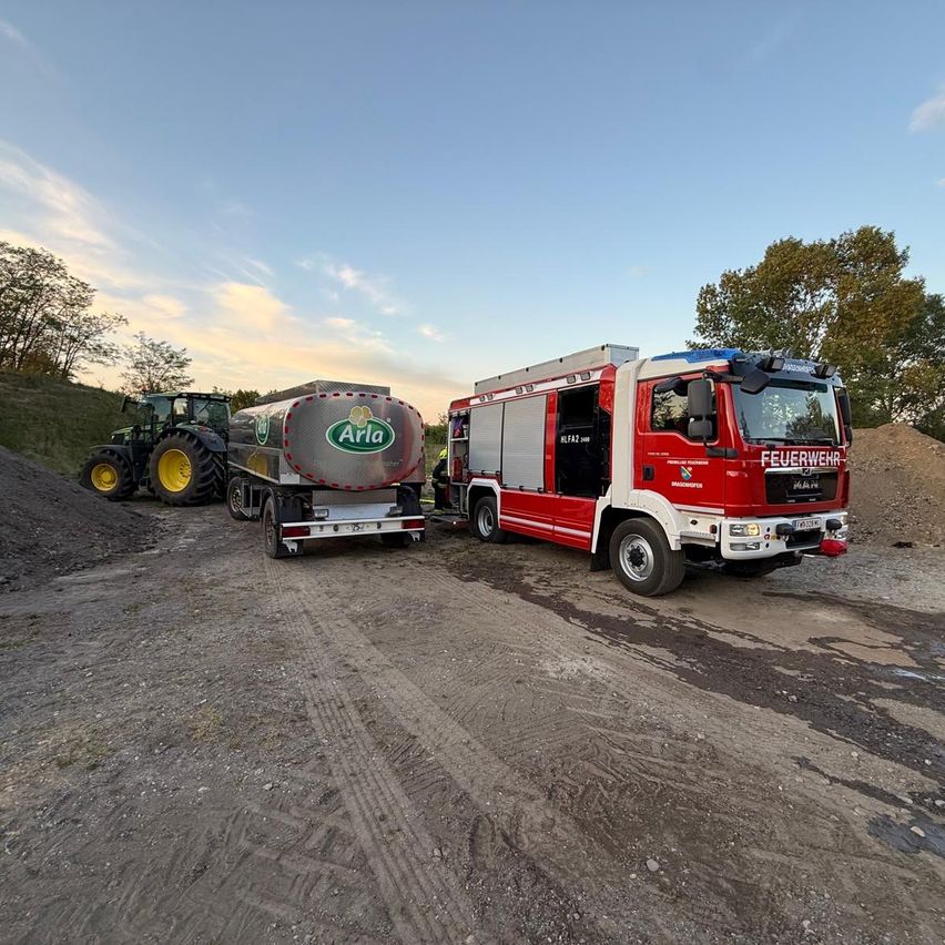 Ein roter Feuerwehrwagen und ein silberner Tankwagen sind auf einer unbefestigten Straße geparkt. Bäume stehen auf beiden Seiten, mit einem klaren Himmel darüber.