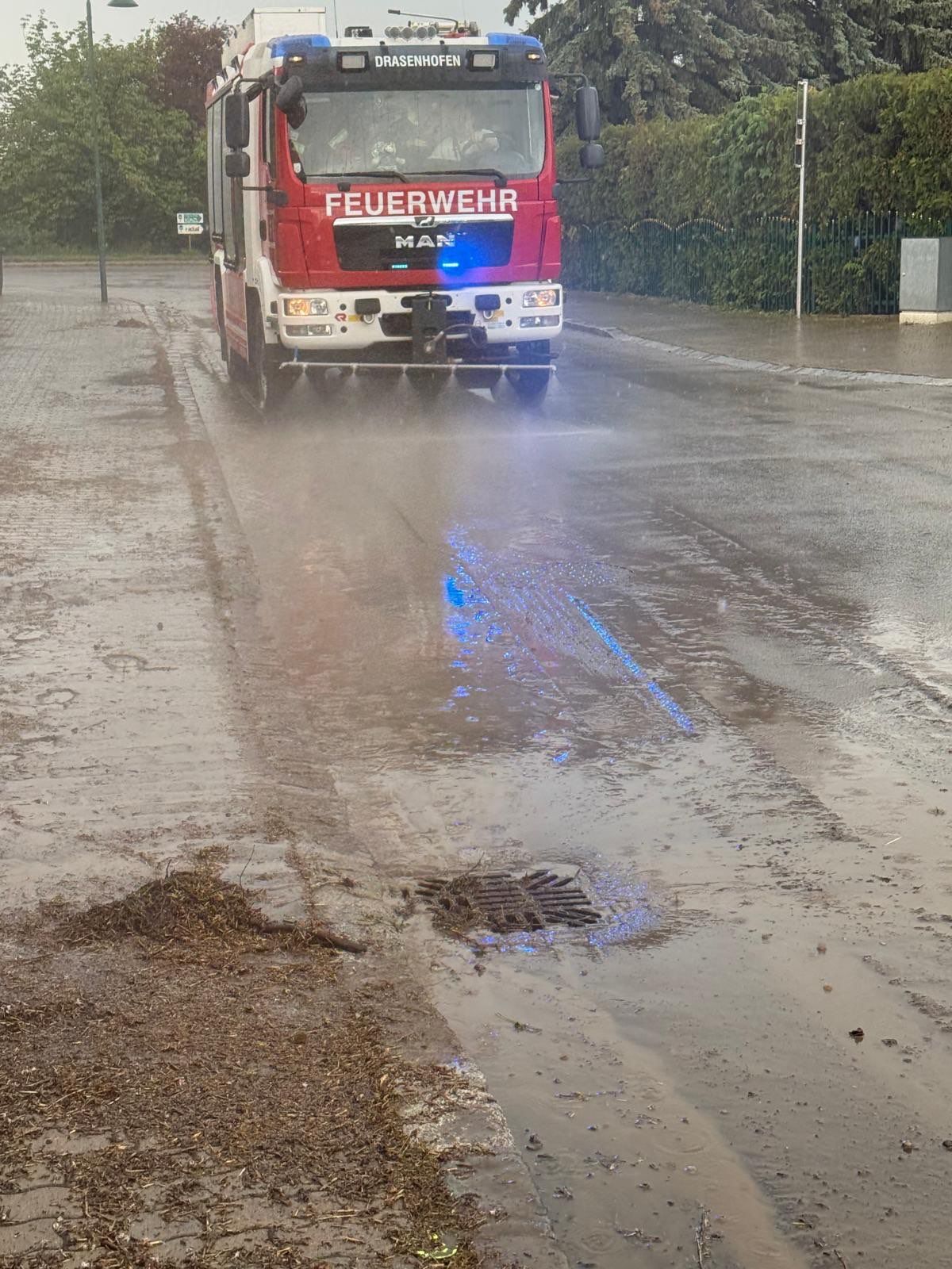 Ein roter Feuerwehrwagen fährt auf einer nassen Straße, seine blauen Lichter blinken. Die Straße ist nass, ein Abflussdeckel ist sichtbar.