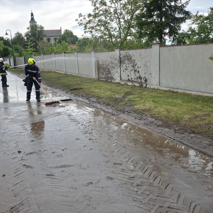 Feuerwehrleute reinigen die verschlammten Straßen nach starkem Regen. Zwei Personen in gelben Helmen kehren die Straße. Eine Betonmauer und Bäume sind im Hintergrund.